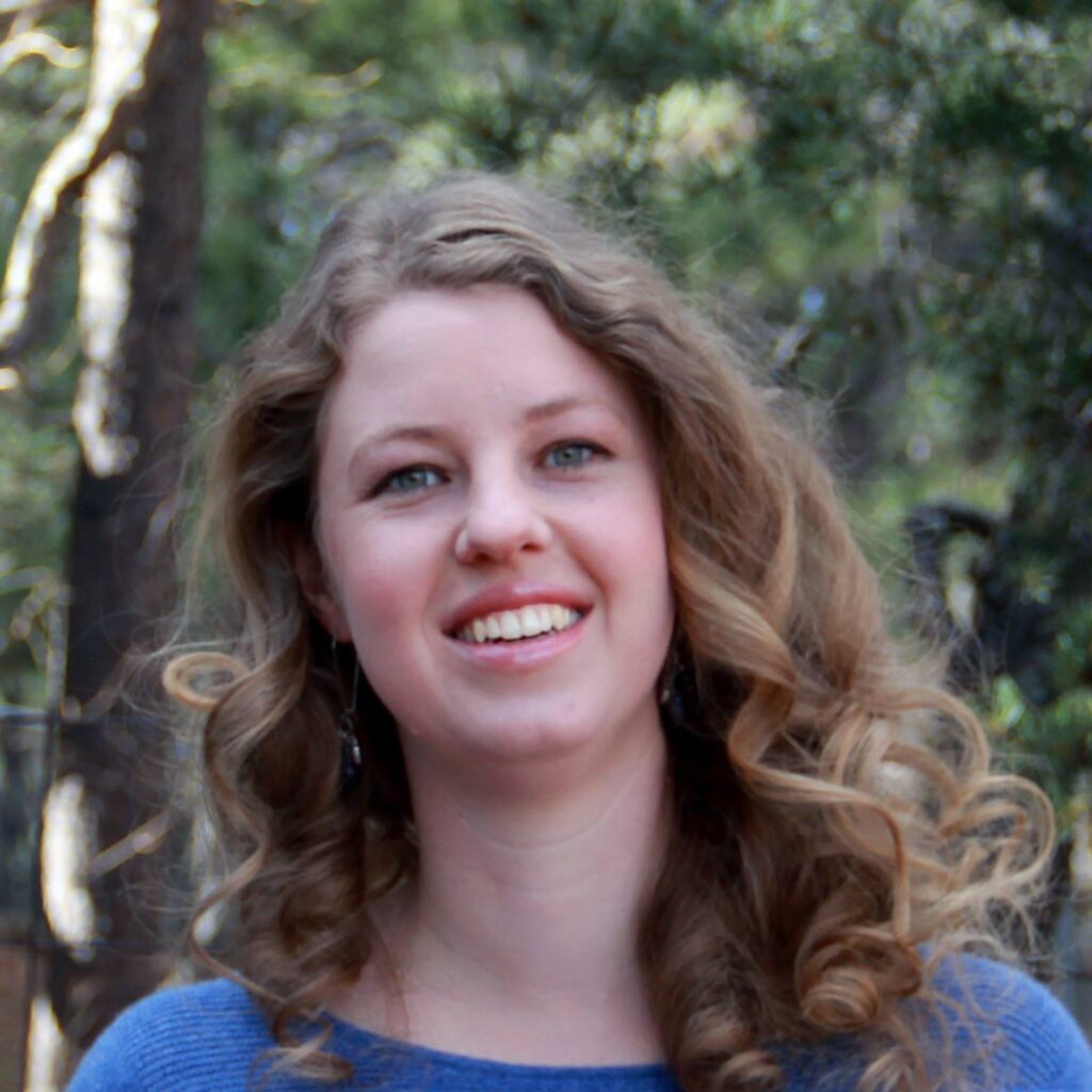 Female smiling with loose curls and a blue shirt in front of foliage